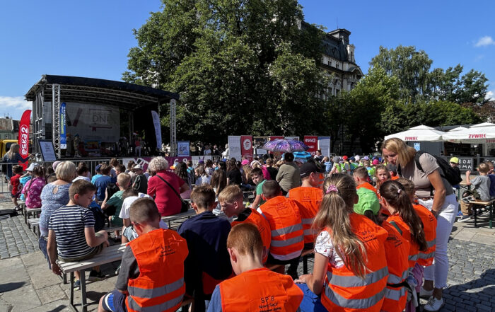 Młodzi ludzie zgromadzeni na dużym placu siedzą skierowani w stronę sceny. Obok widoczny jest fragment zabytkowego ratusza, zielone drzewa oraz błękitne niebo.