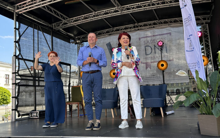 Na scenie po prawej stronie stoi kobieta w białym stroju i kolorowej marynarce, trzyma w ręku mikrofon, uśmiecha się. Obok niej stoi także uśmiechnięty mężczyzna w błękitnej koszuli, także trzymający w rękach mikrofon. Po lewej stronie stoi kobieta w granatowej sukience, macha rękami, tłumaczy rozmowę na język migowy.