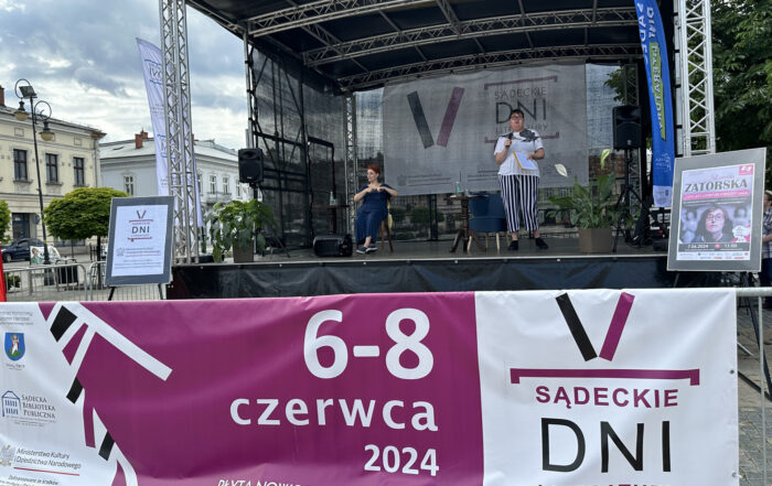 Na scenie stoją dwie kobiety, kobiet, pod sceną umieszczony jest ogromny baner z napisem: 6-8 czerwca 2024, płyta nowosądeckiego rynku, V Sądeckie Dni Literatury.
