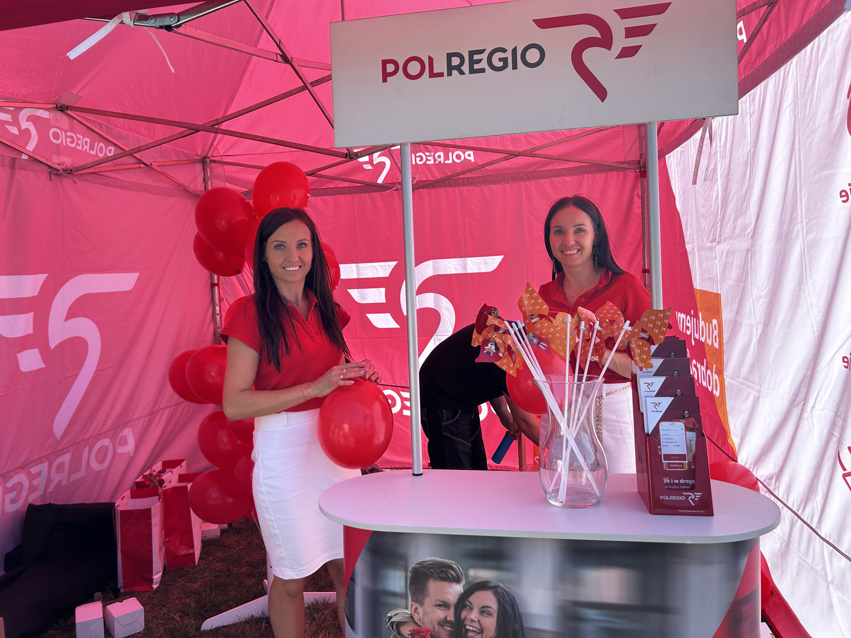 1 (36) Pod czerwono białym namiotem stoją dwie kobiety ubrane w białe spódnice oraz czerwone koszulki. Przed nimi umieszczone jest stoisko z logo Polregio a na półce umieszczone wiatraki oraz ulotki.