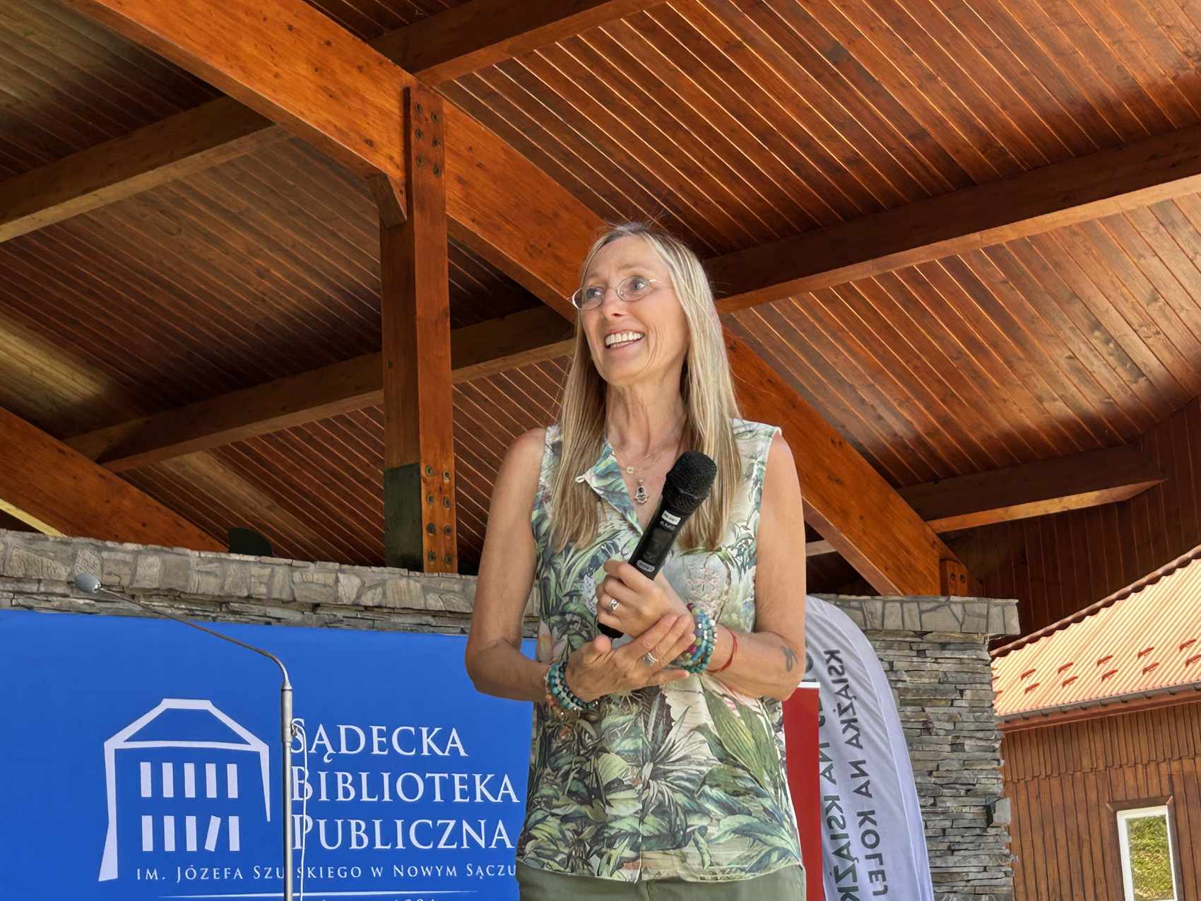 Kobieta ubrana w jasnozieloną bluzkę, widoczna od pasa w górę uśmiecha się. W ręku trzyma mikrofon. Nad nią widoczny jest fragment drewnianego zadaszenia. za nią widnieje na niebieskiej ścianie logo Sądeckiej Biblioteki Publicznej.