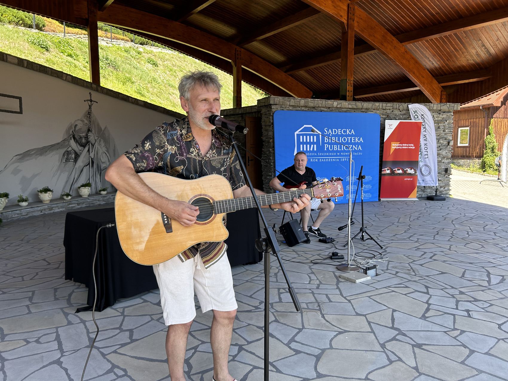 1 (56) Na scenie, pod drewnianym zadaszeniem po lewej stronie stoi mężczyzna z gitarą, śpiewa do mikrofonu. Po prawej stronie siedzi mężczyzna z gitarą. Na nim na niebieskim tle widnieje logo Sądeckiej Biblioteki Publicznej.