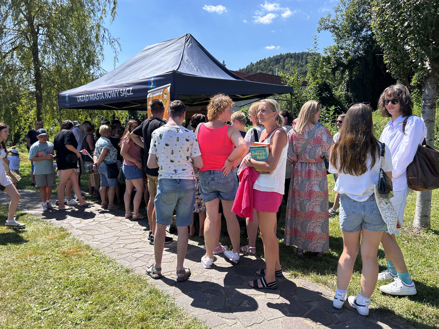 1 (57) Na ścieżce na polance stoi duża grupa ludzi w kolejce do granatowego namiotu z białym napisem Urząd Miasta Nowego Sącza. W tle widoczne są pagórki, drzewa oraz błękitne niebo.