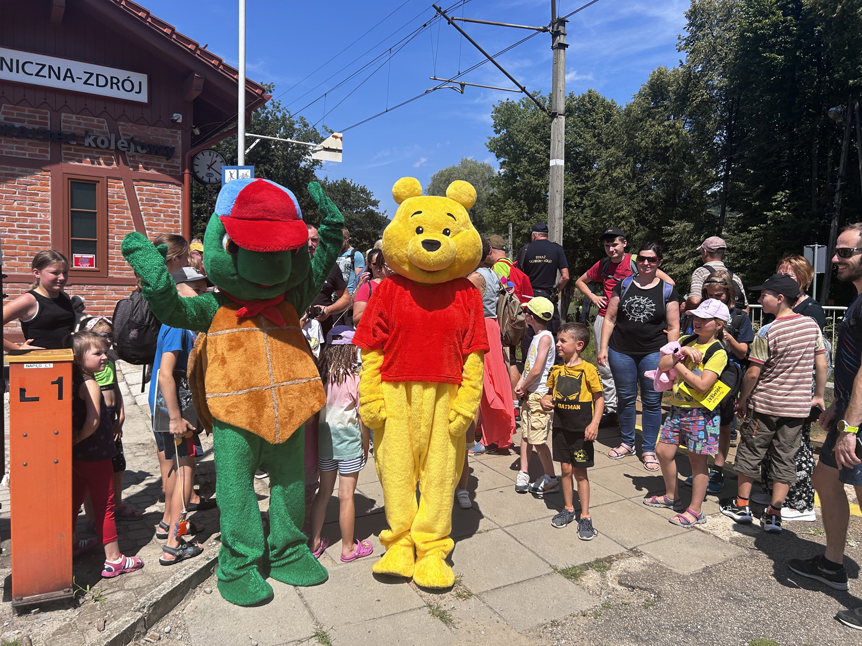 Dworzec kolejowy Piwniczna Zdrój. Duża grupa ludzi stoi na peronie. Przed nimi stoją bajkowe postaci: Żółw franklin i Kubuś Puchatek. W tle widoczne są drzewa oraz błękitne niebo.