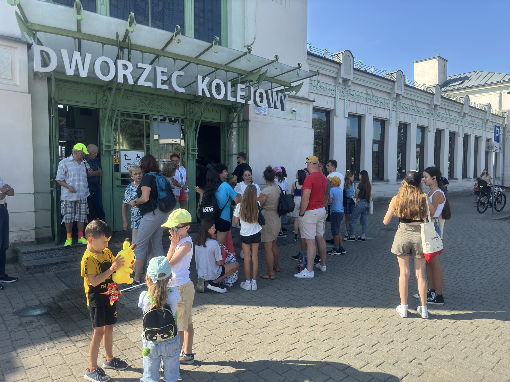 1 (9) Długi, biały budynek z napisem: Dworzec Kolejowy. Grupa ludzi stoi przed wejściem do budynku.