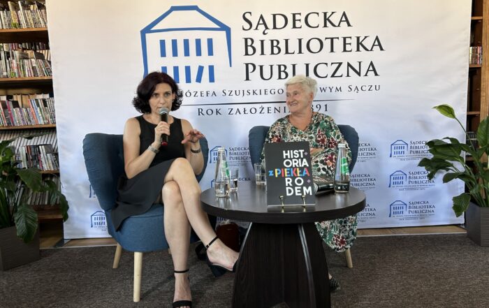 Przy stoliku na fotelach siedzą dwie kobiety. Kobieta po prawej patrzy na kobietę siedzącą po lewej stronie, trzymającą w ręku mikrofon. Za nimi widoczny fragment regału pełnego książek a na tym tle ustawiona ścianka z logo Sądeckiej Biblioteki Publicznej.