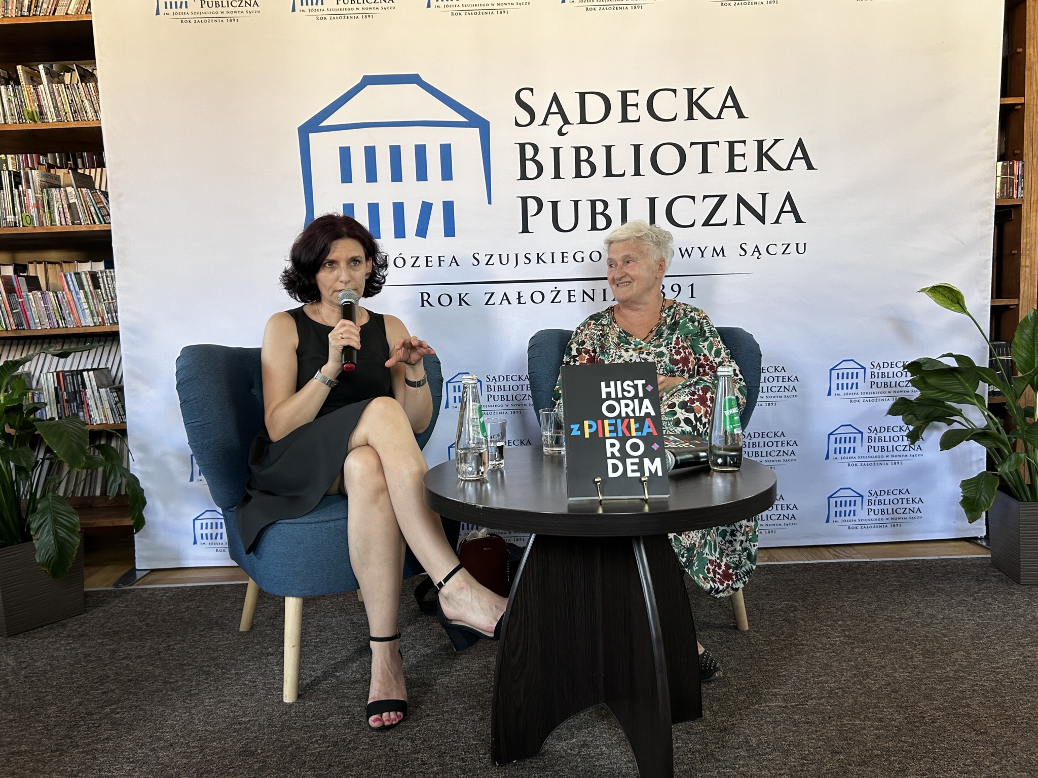 Przy stoliku na fotelach siedzą dwie kobiety. Kobieta po prawej patrzy na kobietę siedzącą po lewej stronie, trzymającą w ręku mikrofon. Za nimi widoczny fragment regału pełnego książek a na tym tle ustawiona ścianka z logo Sądeckiej Biblioteki Publicznej.