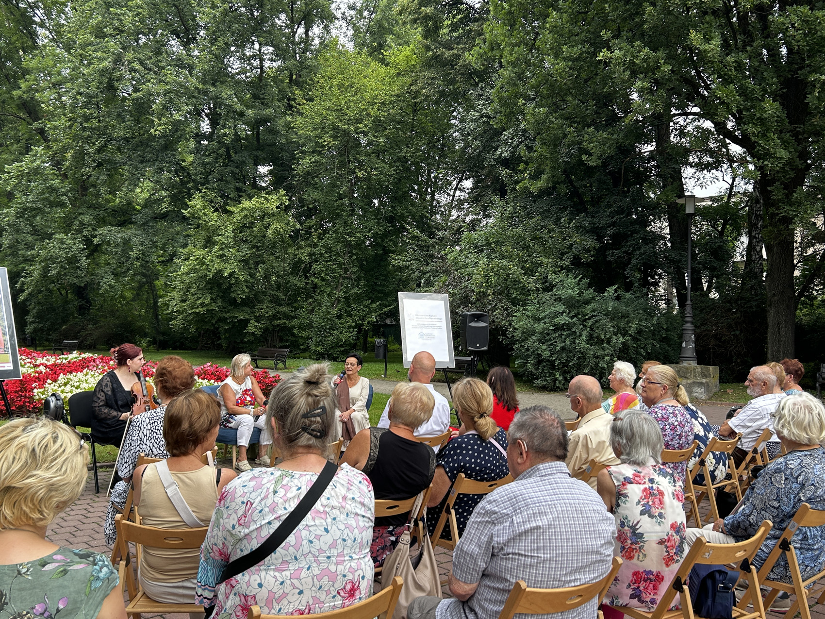 Babiarz Joanna (1) Publiczność zgromadzona w parku siedzi na krzesłach zwrócona w stronę trzech kobiet siedzących przed nimi. Kobieta siedząca po prawej stronę mówi do mikrofonu, kobieta siedząca obok niej przygląda się jej. Siedząca po lewej stronie kobieta trzyma w rękach skrzypce.
