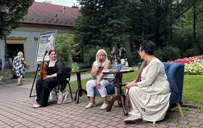 W parku przy stoliku na fotelach siedzą kobiety. Kobieta siedząca po prawej stronie ubrana jest w kremową suknię, przygląda się kobiecie siedzącej po lewej stronie, trzymającej w ręku mikrofon. Po lewej stronie, przy drugim stoliku siedzi ubrana na czarno kobieta trzymająca w ręku skrzypce. W tle widać fragment białego budynku z czerwoną dachówką, ludzi przechodzących alejkami oraz mnóstwo zieleni.