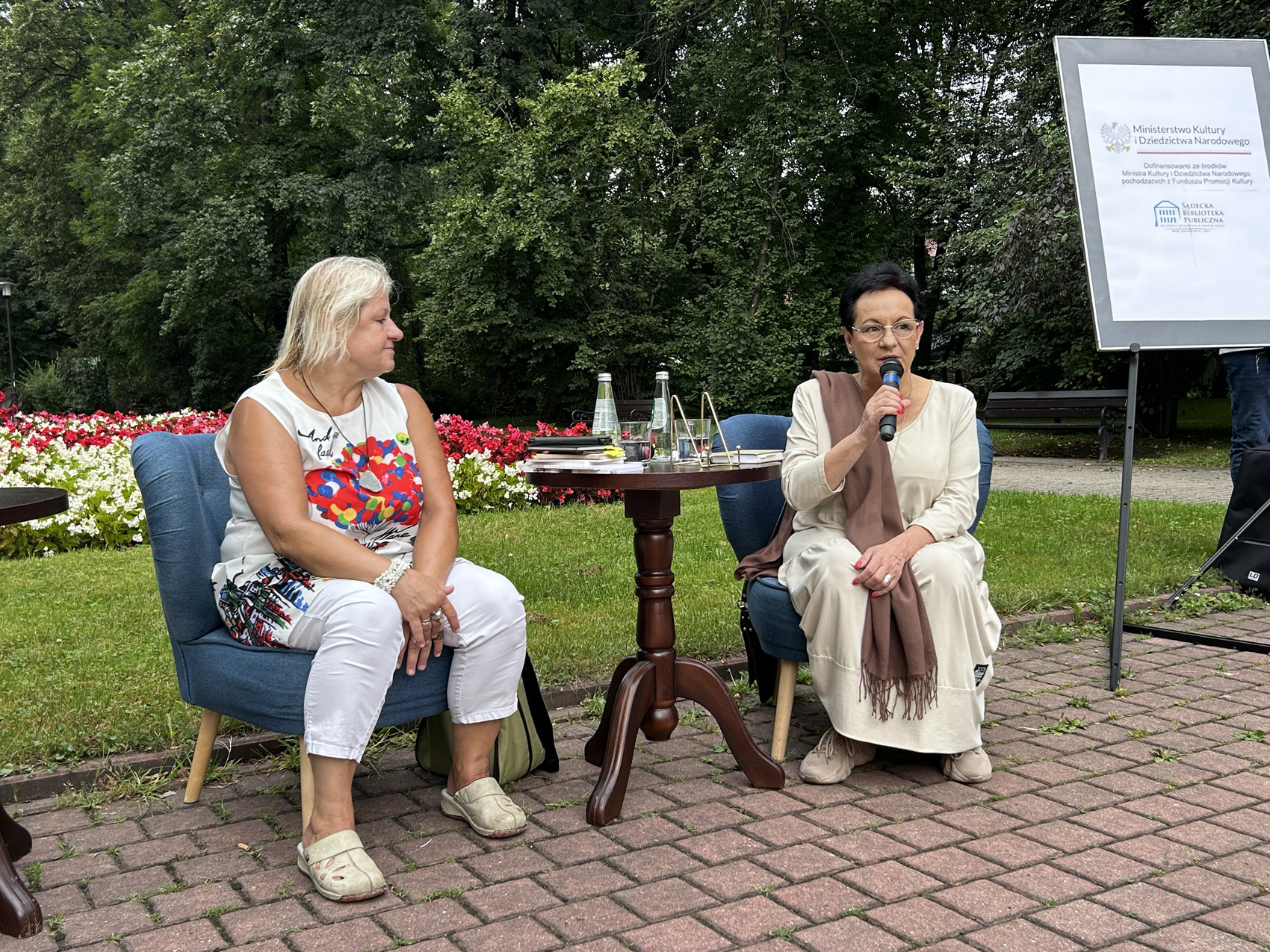 Babiarz Joanna (3) W zielonej przestrzeni miasta przy stoliku na fotelach siedzą dwie kobiety. Kobieta po prawej stronie trzyma w ręku mikrofon, kobieta siedząca po lewej stronie przysłuchuje się. Po prawej stronie ustawiona rama z logo Ministerstwa Kultury i Dziedzictwa narodowego.
