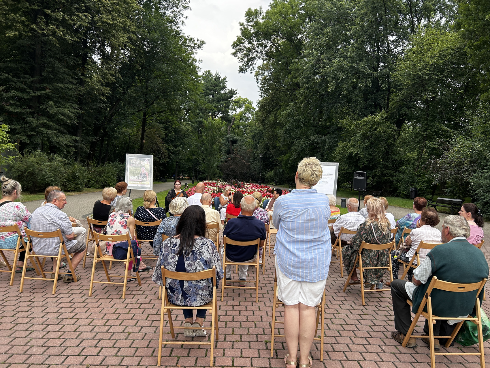 Babiarz Joanna (4) Licznie zgromadzona w parku publiczność siedząca na krzesłach, zwrócona jest w stronę siedzących przed nimi kobiet. Kobieta znajdująca się w środku trzyma w ręku mikrofon. Dookoła widoczne są drzewa oraz alejki.