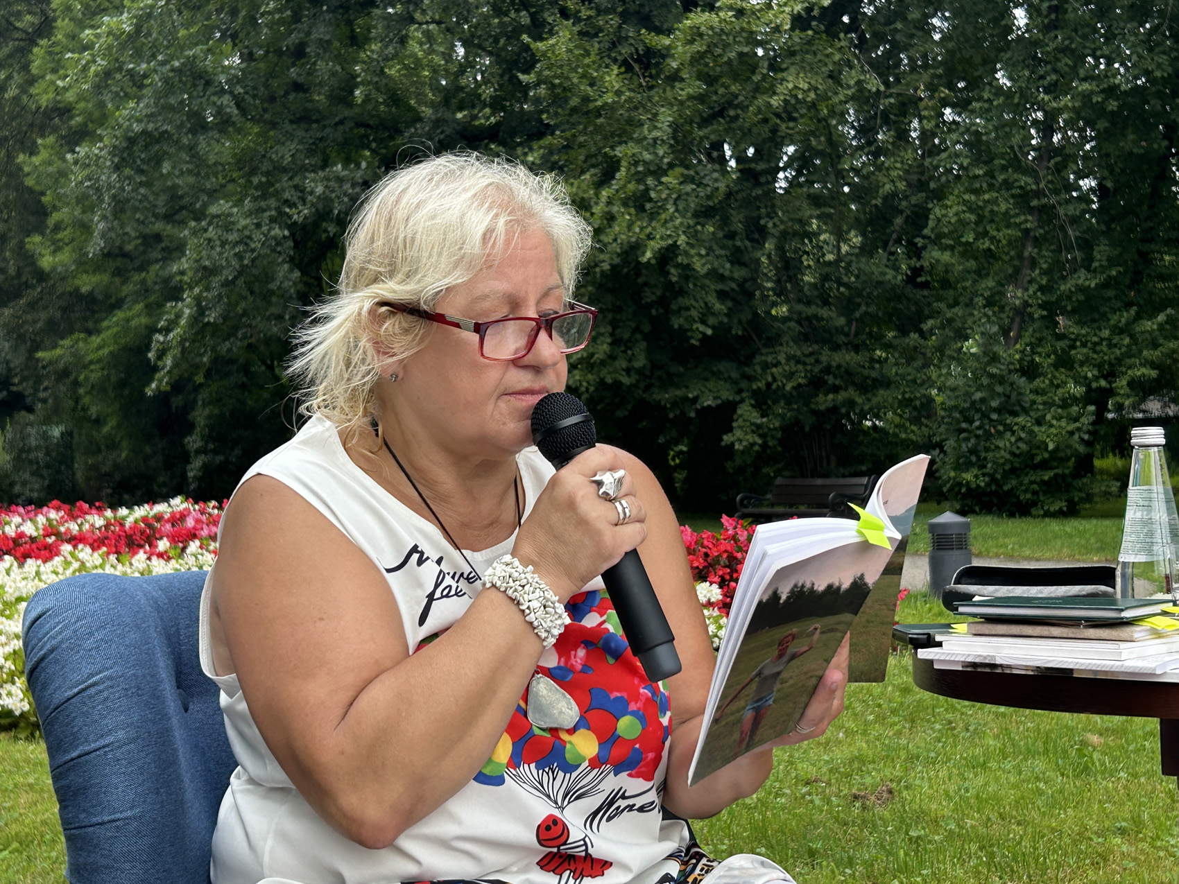 Babiarz Joanna (5) Siedząca na fotelu kobieta w jednej ręce trzyma mikrofon, w drugiej książkę. Przy niej widoczny jest fragment stolika z książkami oraz szklana butelka. Dookoła widoczne są drzewa oraz czarno białe kwiaty.