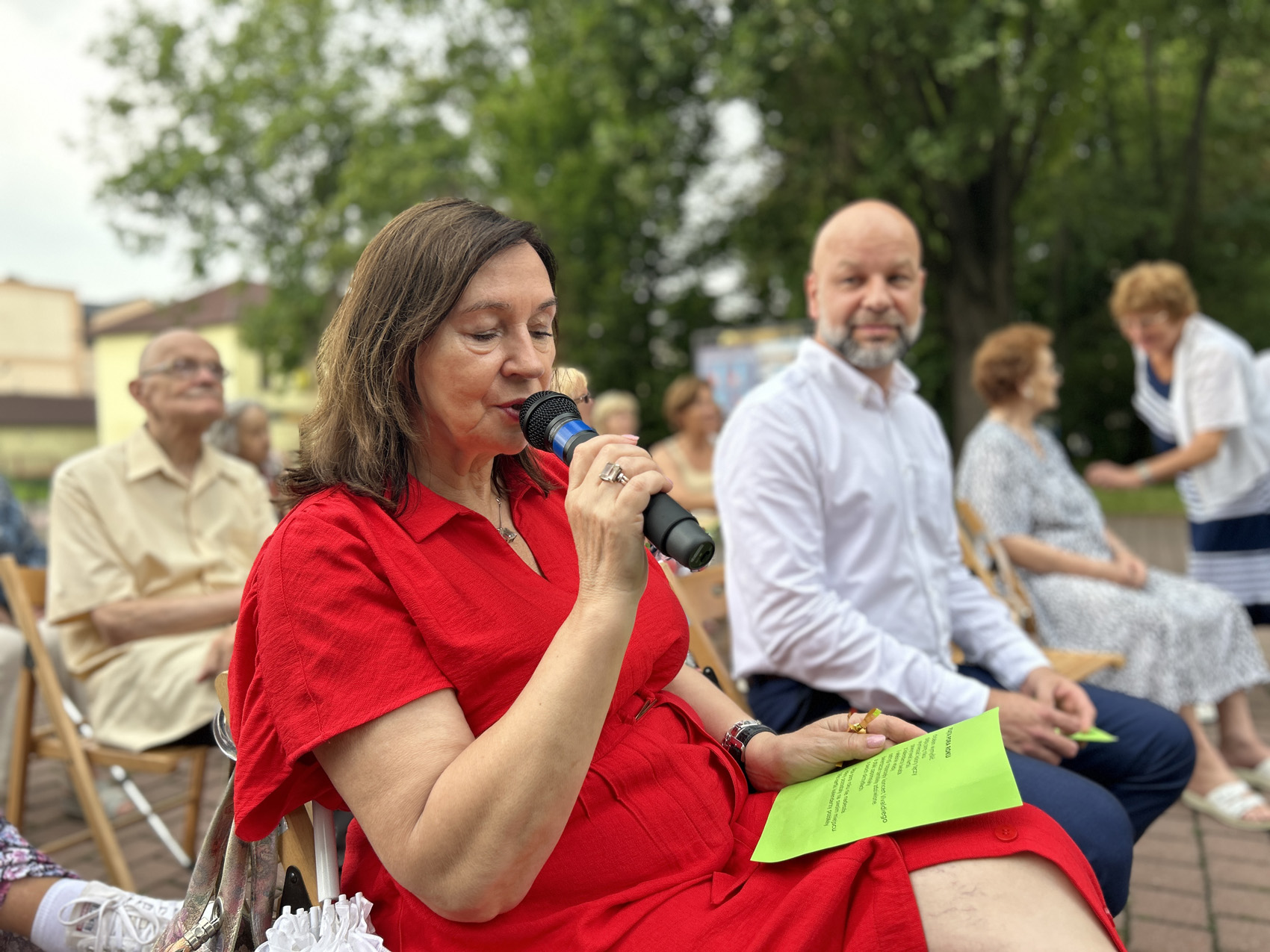 Babiarz Joanna (6) Kobieta w czerwonej sukience trzyma w ręku mikrofon, drugą ręką przytrzymuje zieloną karteczkę, leżącą na jej kolanach. W tle widoczne są osoby siedzące obok na krzesłach.