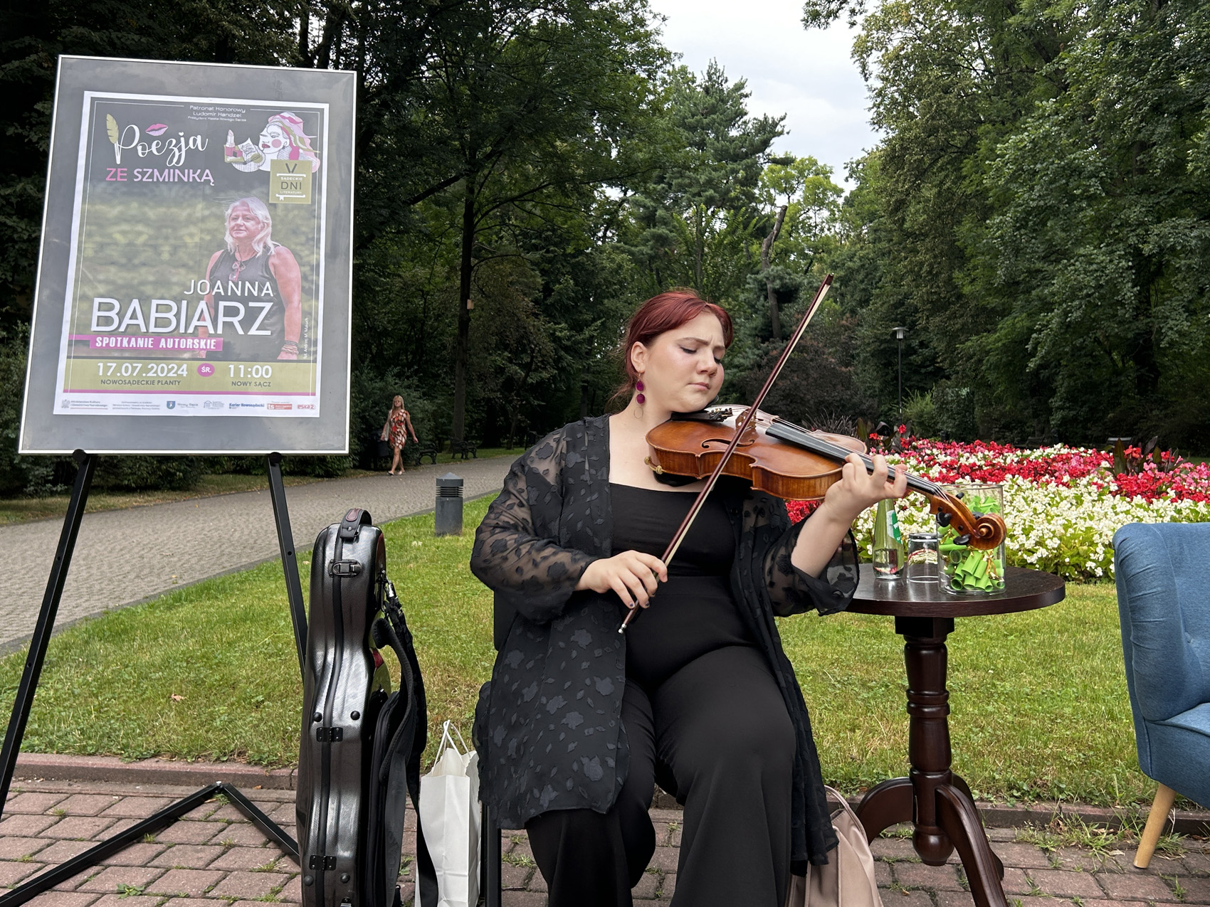 Babiarz Joanna (8) Przy stoliku siedzi ubrana na czarno kobieta grająca na skrzypcach. Obok niej po prawej stronie ustawiony jest stolik z kolorowymi karteczkami umieszczonymi w szklanym słoju oraz szklana butelka. Po lewej stronie umieszczona jest sztaluga z wizerunkiem kobiety na napisem: Poezja ze szminką. Joanna Babiarz. W tle widoczne są drzewa, czerwono białe kwiaty oraz alejki.