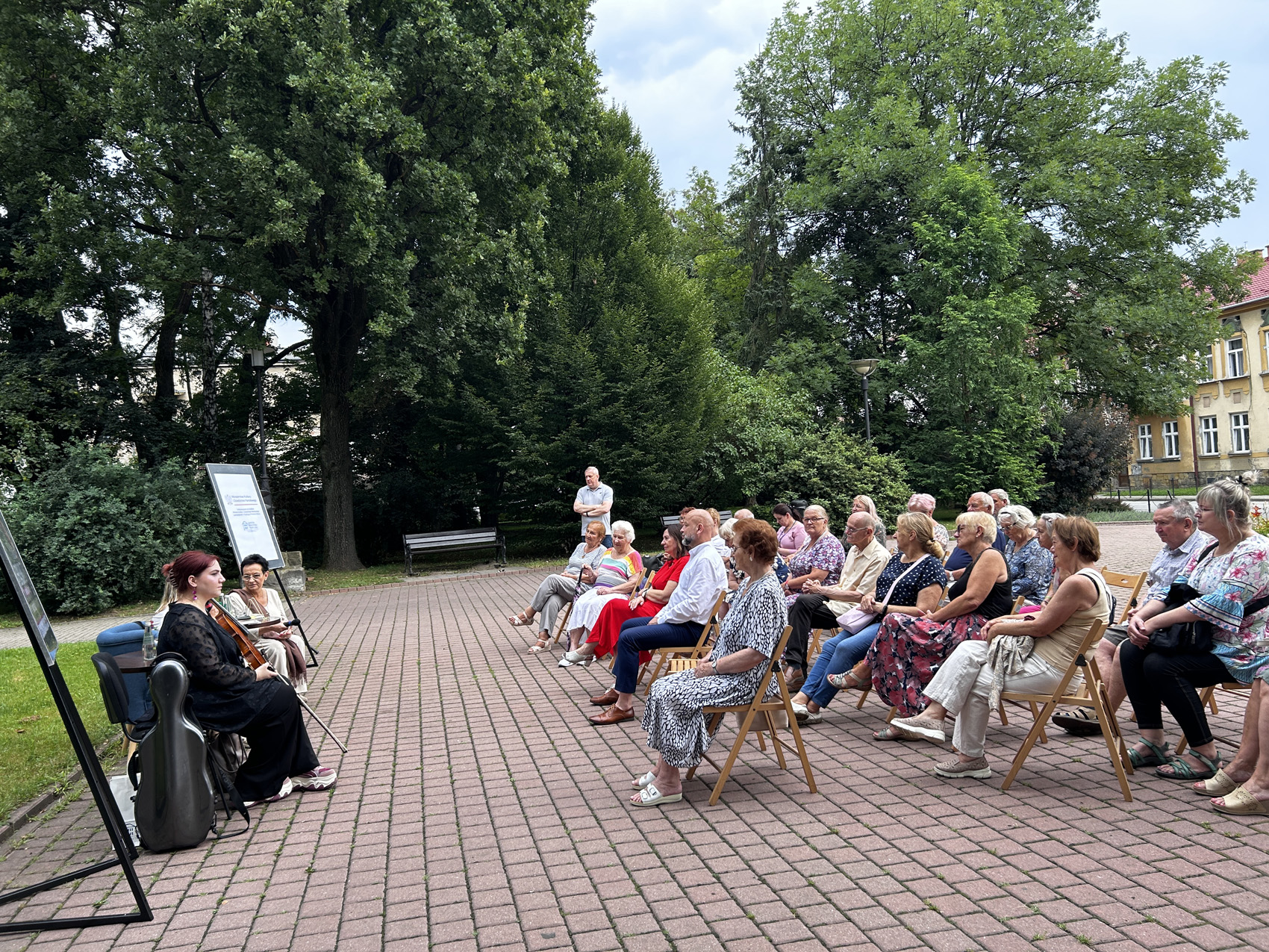 Babiarz Joanna (9) Na chodniku wśród drzew po lewej stronie siedzą trzy kobiety. Jedna z nich trzyma w rękach skrzypce. Po prawej stronie na krzesłach siedzi zgromadzona publiczność, przyglądająca się kobietom.