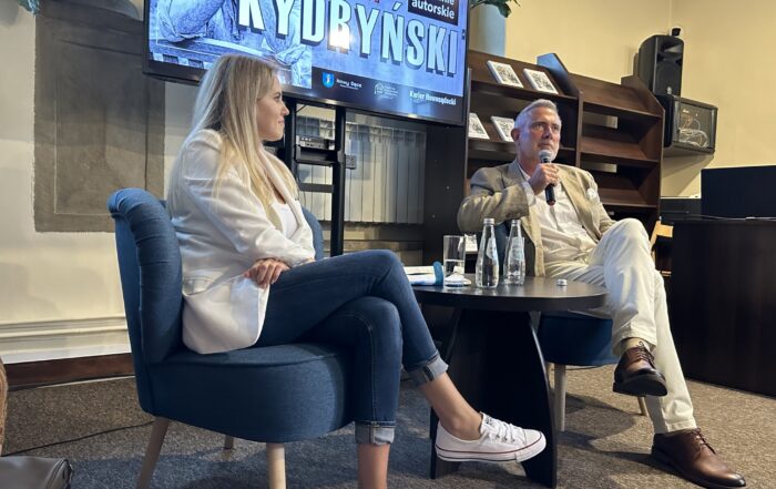 Kobieta i mężczyzna siedzą przy stoliku, na którym umieszczone są szklane butelki. Mężczyzna siedzący po prawej stronie mówi do mikrofonu, kobieta siedząca obok przygląda się mu. Za nimi widnieje ekran z czerwono białym napisem: Marcin Kydryński.