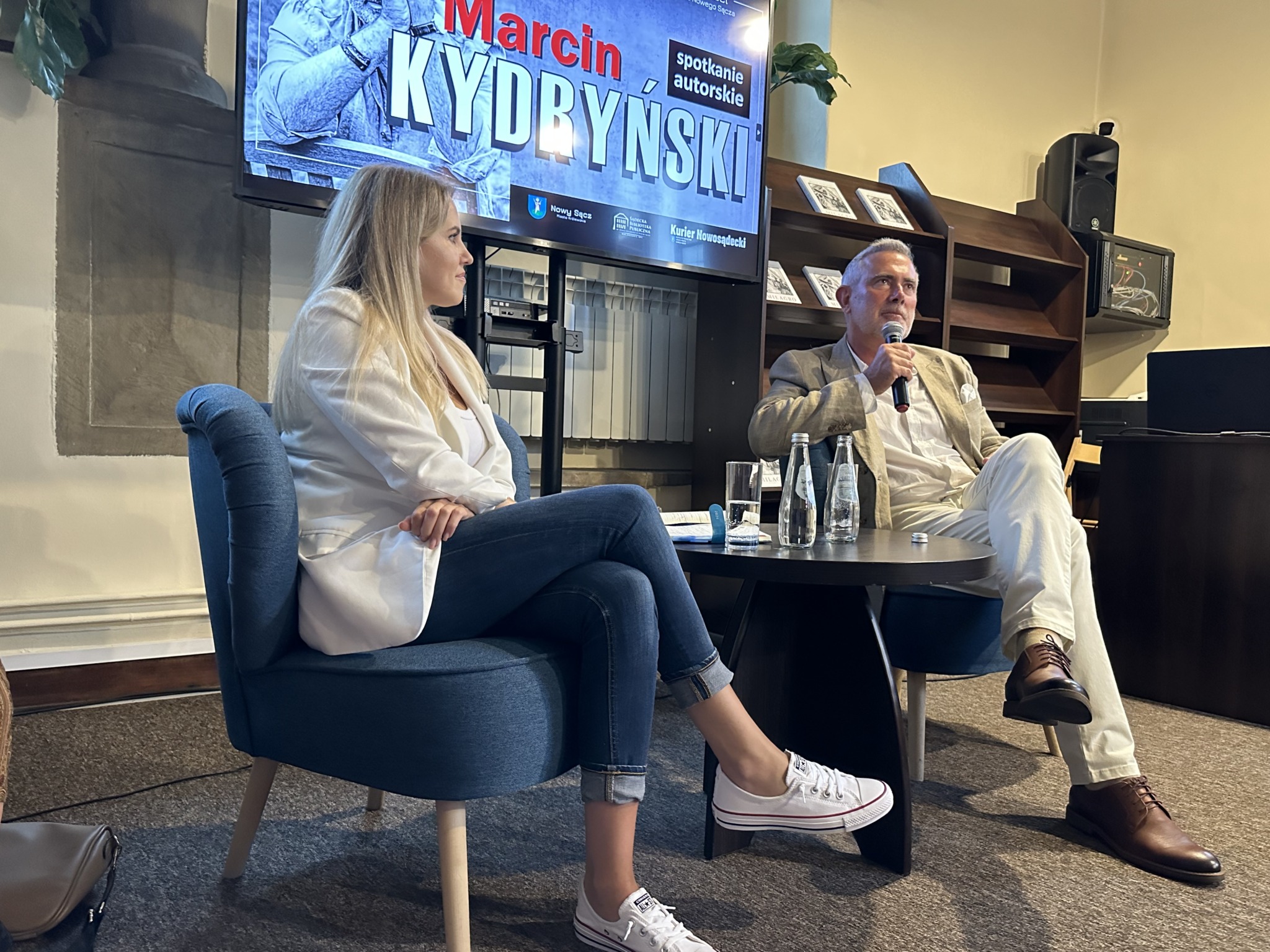 Kobieta i mężczyzna siedzą przy stoliku, na którym umieszczone są szklane butelki. Mężczyzna siedzący po prawej stronie mówi do mikrofonu, kobieta siedząca obok przygląda się mu. Za nimi widnieje ekran z czerwono białym napisem: Marcin Kydryński.