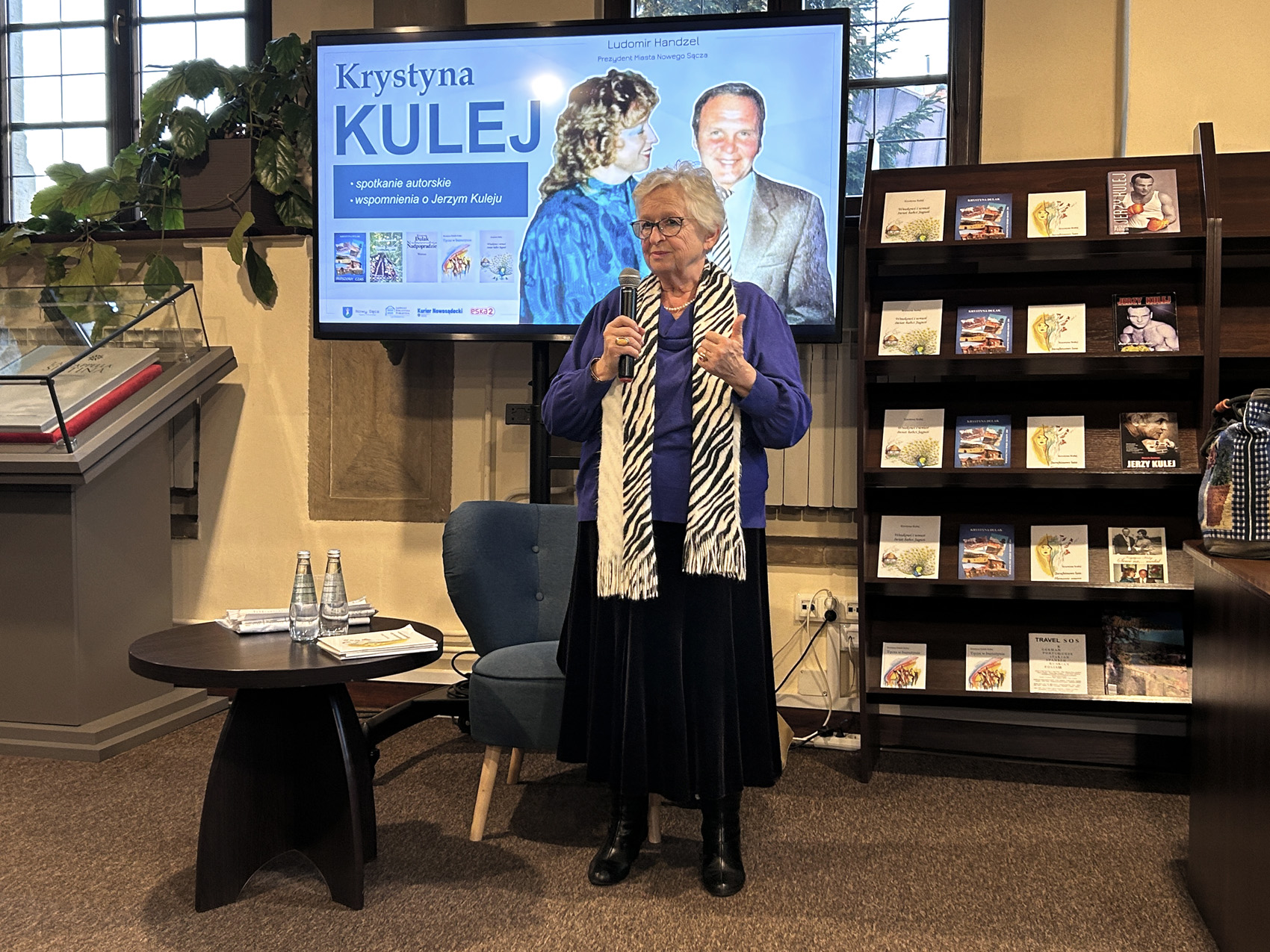 Krrystyna Kulej (6) Kobieta z mikrofonem stoi obok dużego regału z gazetami. Za nią jest duży ekran z prezentacją o Krystynie Kulej.