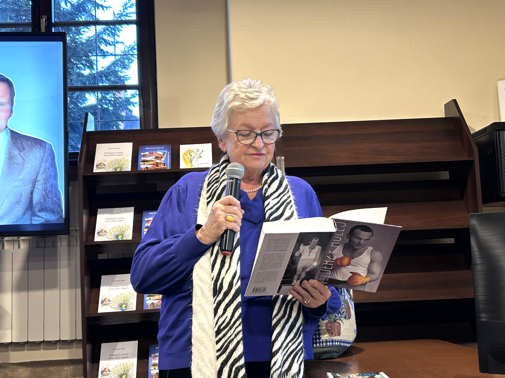 Krystyna Kulej (1) Starsza kobieta czyta książkę, w jednej ręce trzyma mikrofon, a w drugiej otwartą książkę.