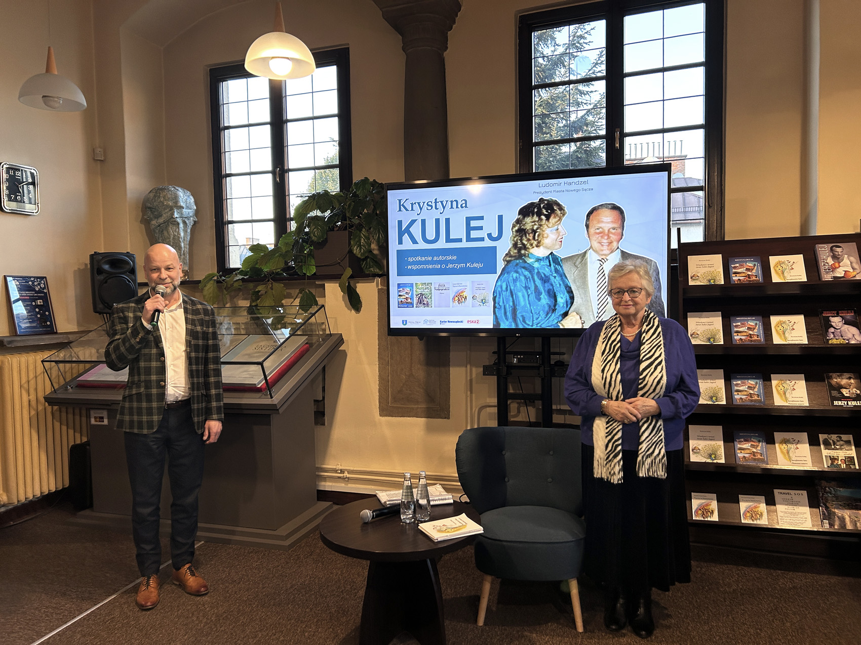 Krystyna Kulej (2) Dyrektor biblioteki w Nowym Sączu trzyma przy twarzy mikrofon. Obok niego stoi starsza kobieta. Za nimi znajduje się gablota z książkami i duży ekran.