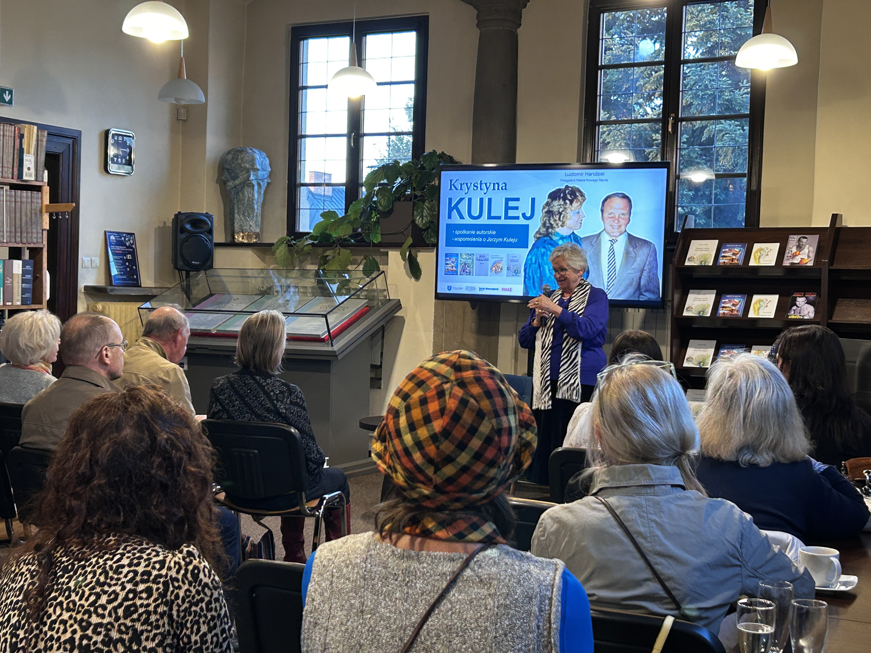 Krystyna Kulej (3) Kobieta z mikrofonem stoi przed dużą grupą ludzi, która siedzi na krzesłach. Za nią jest duży ekran z prezentacją o Krystynie Kulej.