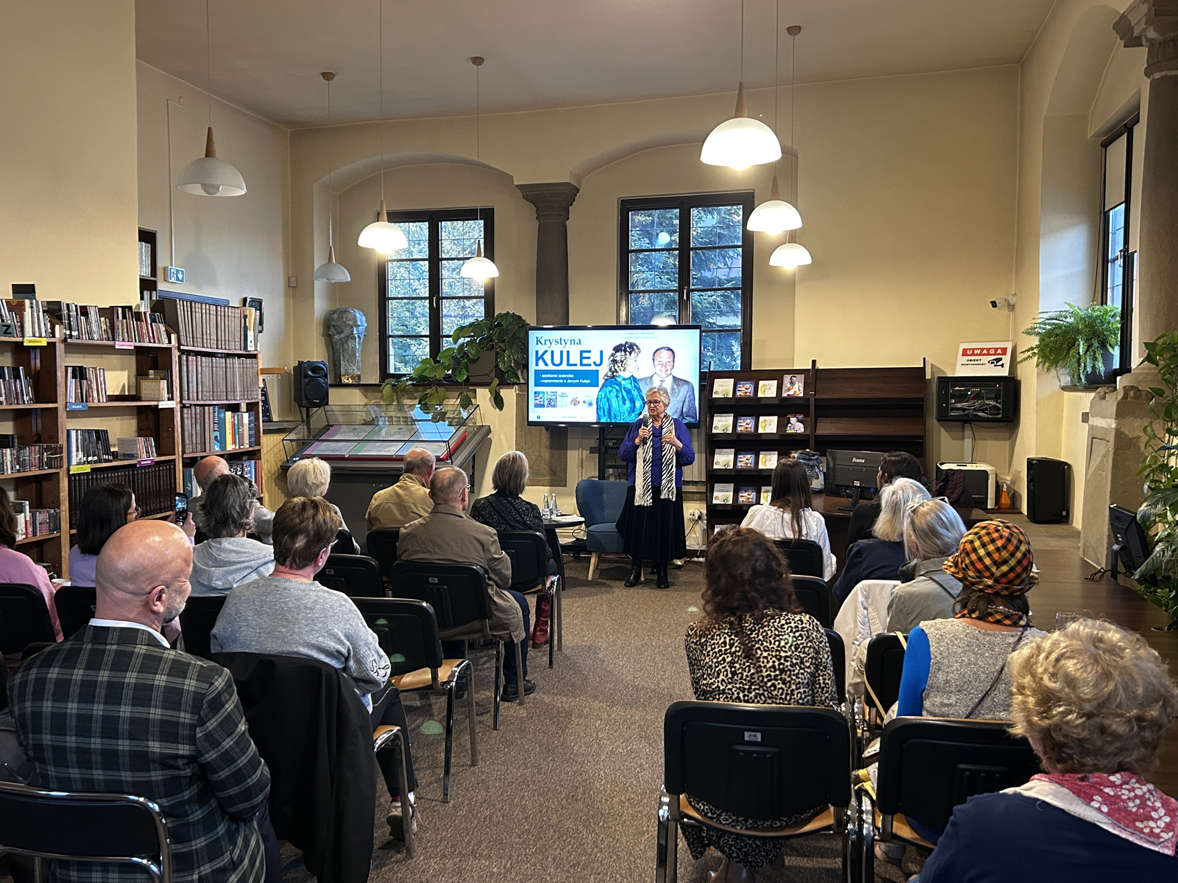 Krystyna Kulej (4) Kobieta z mikrofonem stoi przed dużą grupą ludzi, która siedzi na krzesłach. Za nią jest duży ekran z prezentacją o Krystynie Kulej.