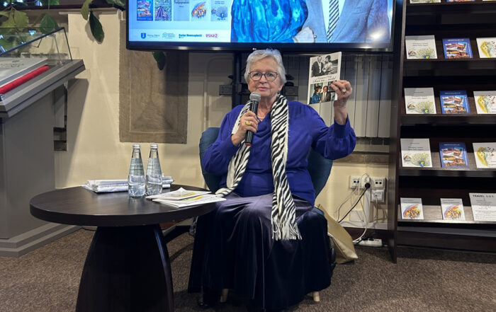 Starsza kobieta siedzi na krześle przy stoliku i mówi do mikrofonu, w drugiej ręce trzyma książkę.