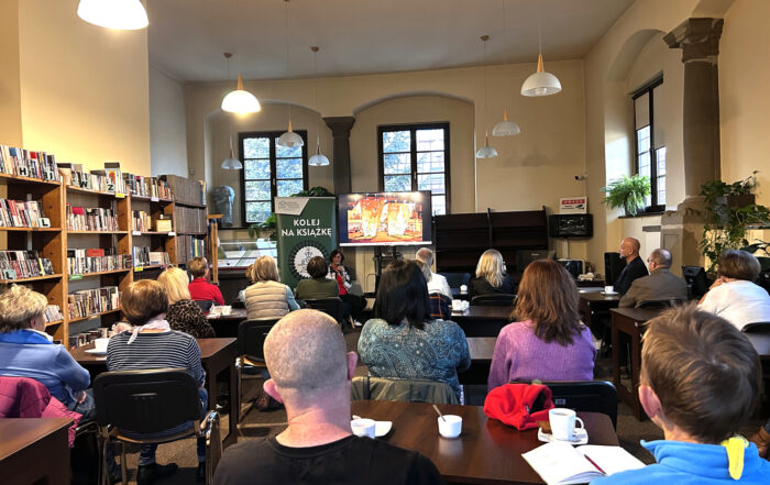 Sala pełna ludzi, które siedzą przy stolikach i słuchają wykładu kobiety. Spotkanie z serii "Kolej na książkę, książka na kolej"