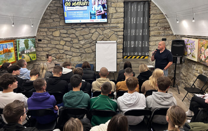 Grupa uczniów podczas spotkania, która słucha mężczyzny z mikrofonem.