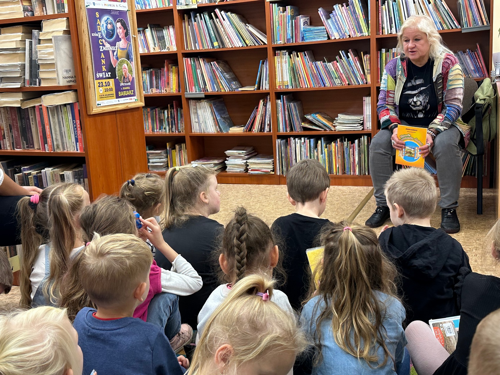 Sądeczaniki_babiarz_1 Starsza kobieta trzyma w rękach książkę i mówi do grupy dzieci, które siedzą na podłodze, na tle pełnych regałów z książkami.
