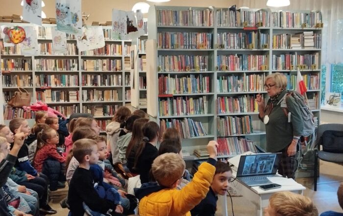 Kobieta z plecakiem, do którego włożona jest polska flaga stoi przed grupą dzieci i opowiada. W tle na ścianie duży regał z książkami.