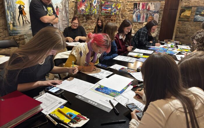 Na zdjęciu jedenaście osób. Pośrodku duży czarny stół. Na blacie stołu leżą bloki techniczne, kredki i cienkopisy. Po lewej stronie rysują cztery osoby. Dziewczyna siedząca przy stole po prawej stronie o włosach w kolorze brązowym patrzy w telefon. Mężczyzna stojący z tyłu patrzy na naszkicowany komiks