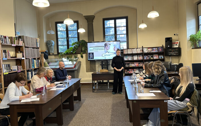 Dwa stoły za którymi siedzi młodzież i seniorzy. Z przodu stoi kobieta.