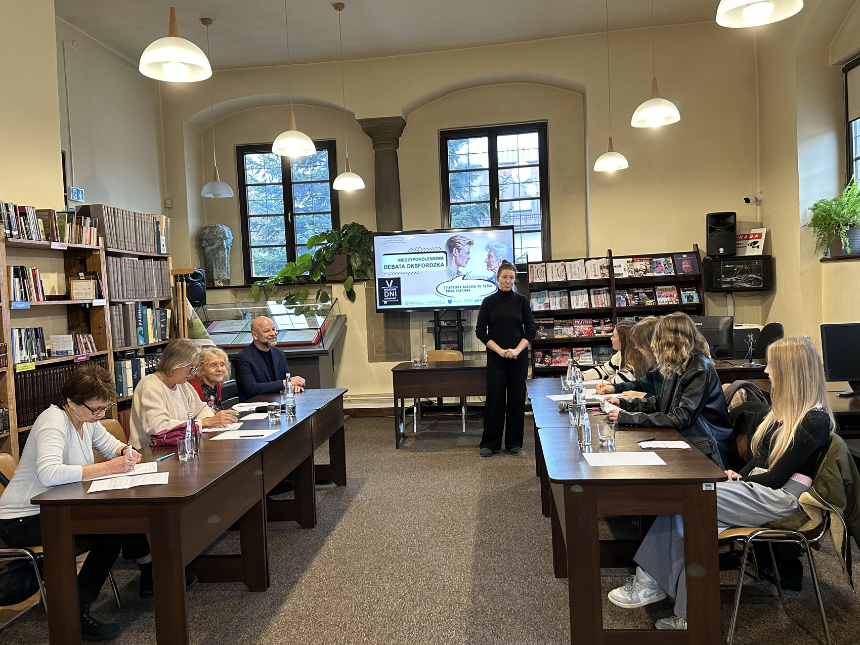 Dwa stoły za którymi siedzi młodzież i seniorzy. Z przodu stoi kobieta.