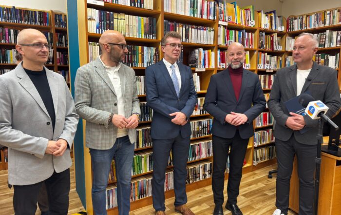 Prezydent, dyrektor biblioteki oraz inni ludzie stoją na tle regałów z książkami.