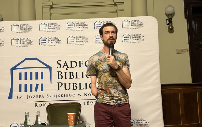 Mężczyzna stoi na tle roll-up i mówi do mikrofonu.