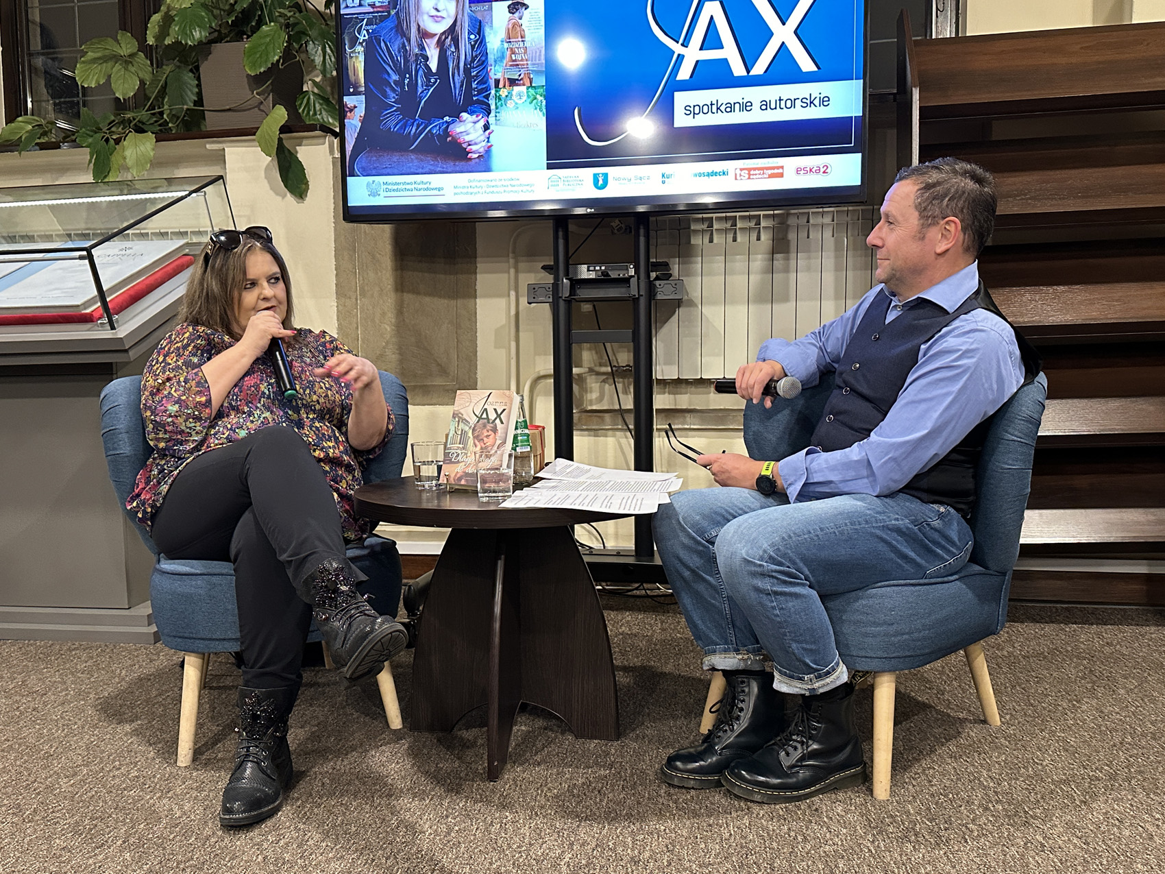 Dwie osoby siedzą na krzesłach przy stoliku i rozmawiają.