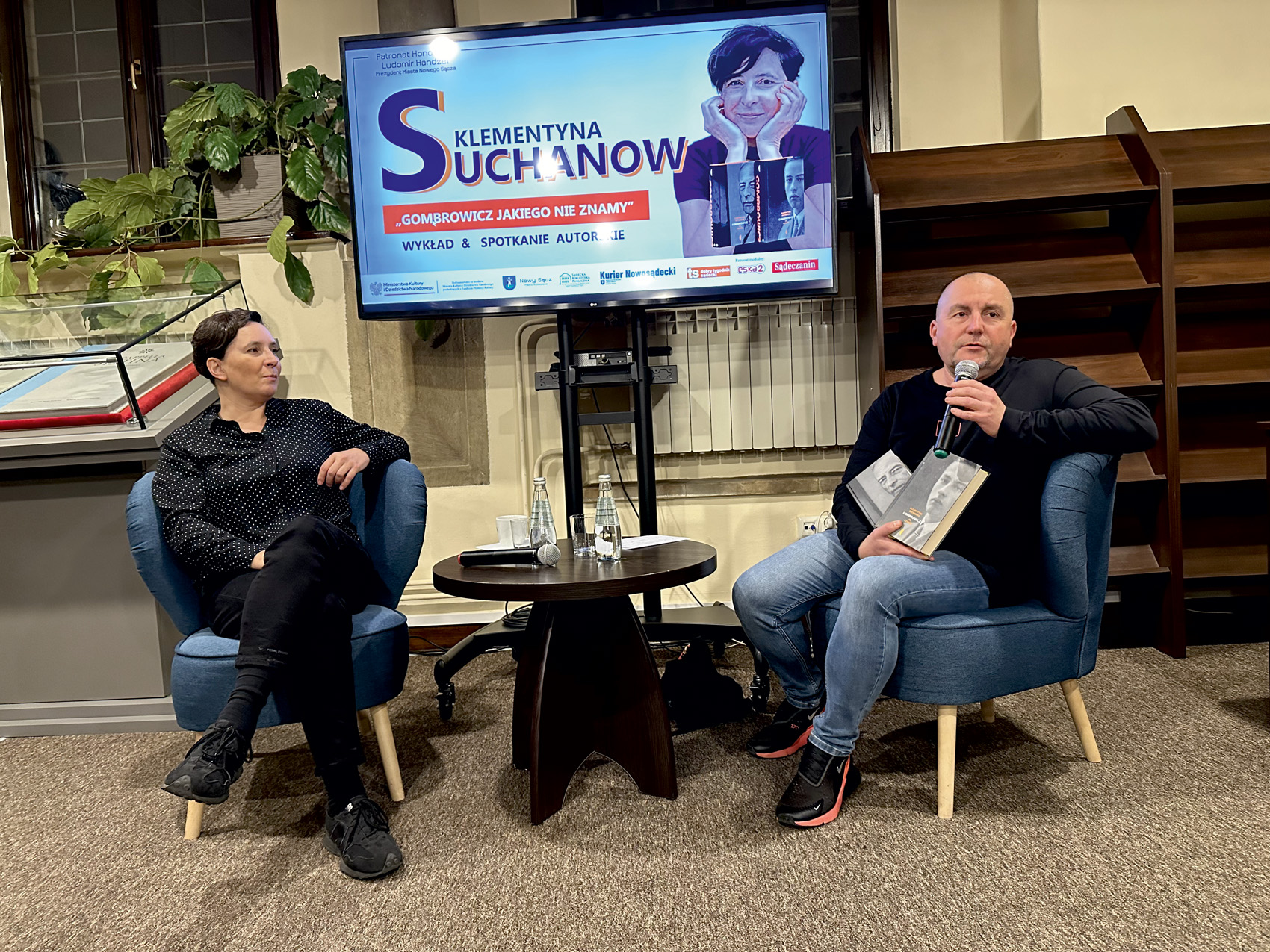 Kobieta i mężczyzna siedzą na krzesłach i opowiadają o książce. Pomiędzy nimi jest ekran.