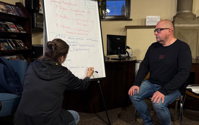 Kobieta kuca i pisze po białej tablicy, a mężczyzna siedzi na krześle i patrzy na zapiski kobiety.