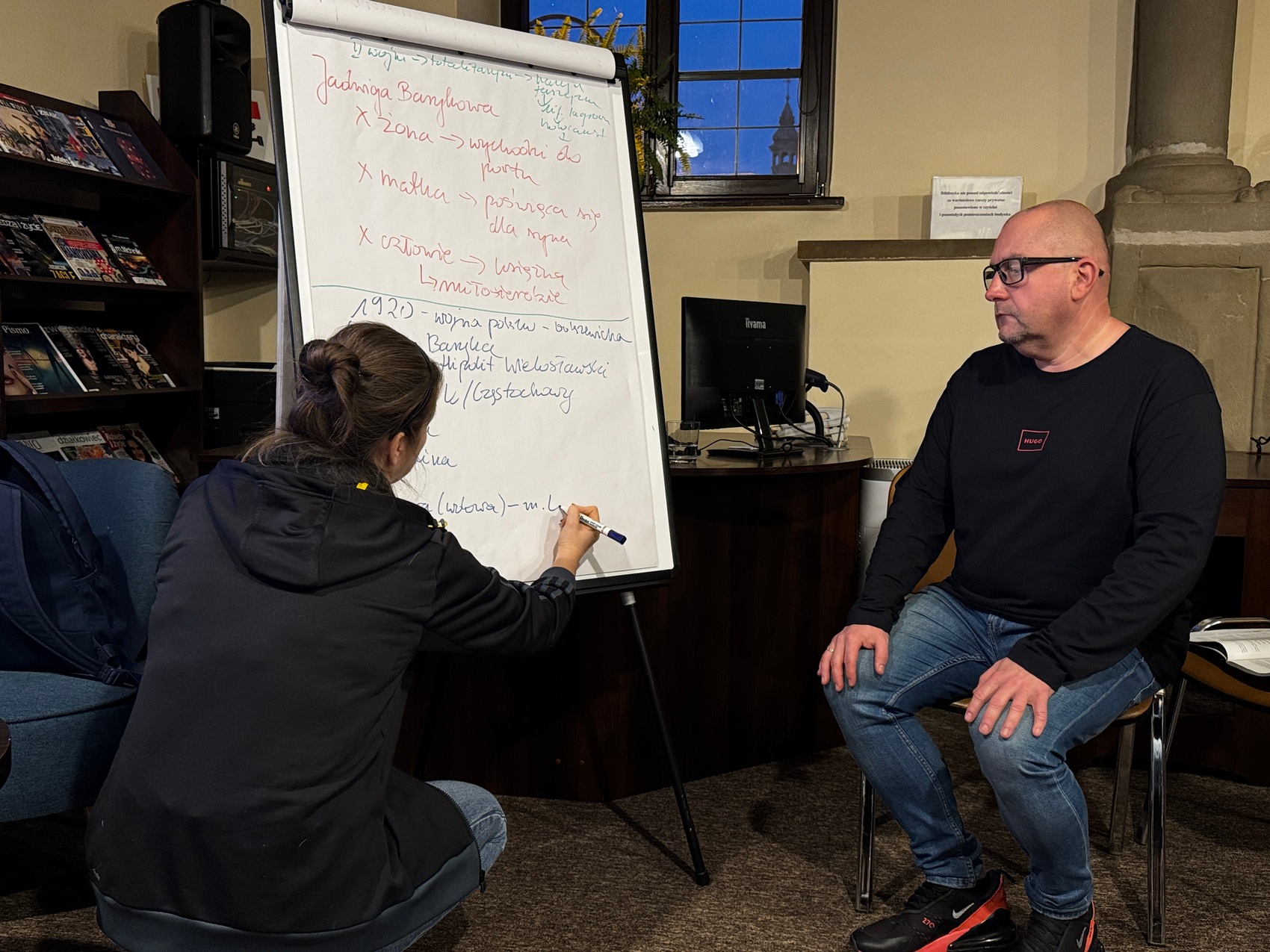 Kobieta kuca i pisze po białej tablicy, a mężczyzna siedzi na krześle i patrzy na zapiski kobiety.