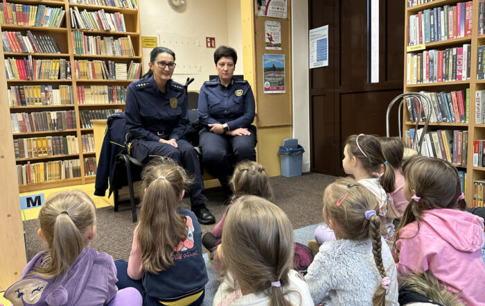 Grupa dzieci siedzi na podłodze i patrzy na siedzące przed nimi dwie policjantki.