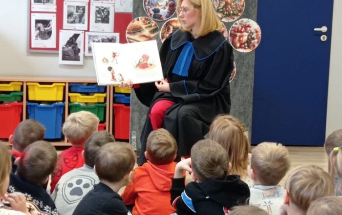 Sala dla dzieci. Na podłodze na zielonej macie siedzi grupa małych dzieci. Patrzą przed siebie. Przed nimi na krześle siedzi kobieta w długiej czarnej todze. Trzyma w rękach otwartą książkę. Za nią na ścianie naklejone są świąteczne grafiki w kształt choinki, obok ściany półki z kolorowymi plastikowymi pudełkami.