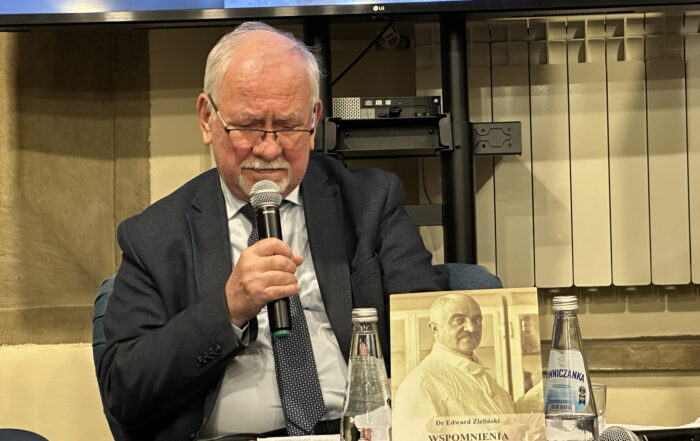 Starszy siwy mężczyzna z wąsami i okularami siedzi na fotelu. Ma sobie garnitur. Trzyma mikrofon przy twarzy. Przed nim jest stolik. Na stoliku jest książka, dwie szklane butelki wody i dwa mikrofony. Za mężczyzna znajduje się stelaż metalowego czarnego ekranu.