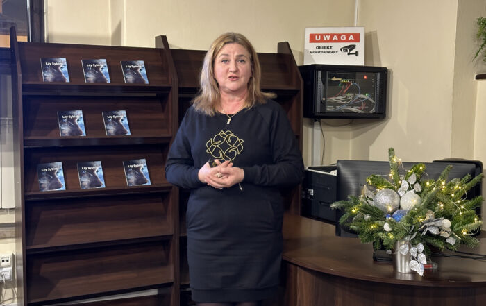 Kobieta w jasnych włosach do ramion i czarnej krótkiej sukience stoi na tle regału z książkami. Obok stoi lada. Na ladzie bożonarodzeniowy stroik.