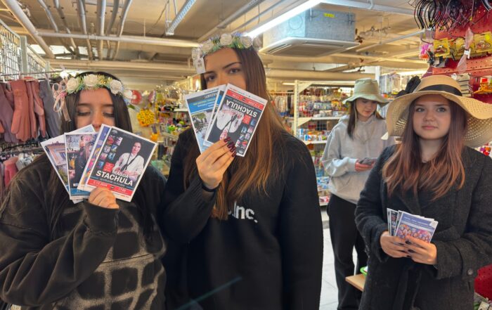 Cztery dziewczyny z ulotkami w ręce. Dwie z nich mają wianki na głowie, a dwie kapelusze. W tle regały sklepowe z kolorowymi przedmiotami.