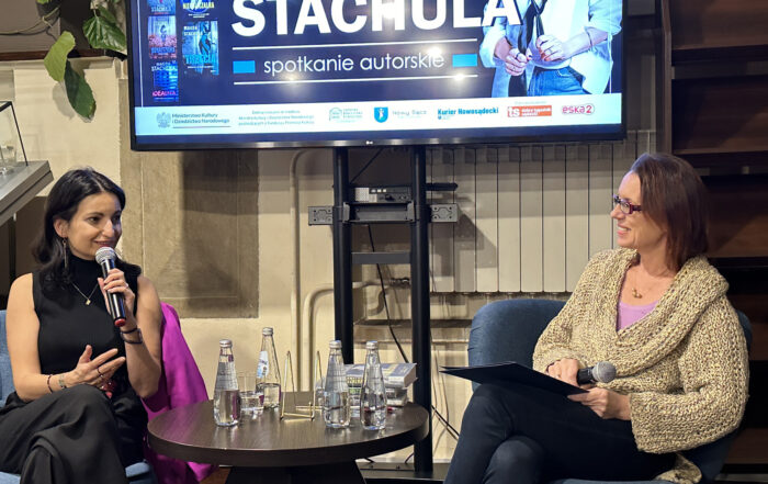 Dwie kobiety siedzą przy niskim okrągłym stoliku. Na stoliku książki i szklane butelki wody. Kobieta po lewej ma czarny strój. Trzyma w rękach mikrofon. Uśmiecha się. Patrzy na drugą kobietę. Kobieta po prawej uśmiecha się. Trzyma w jednej ręce teczkę, a w drugiej mikrofon. Ma na sobie gruby jasny sweter i ciemne spodnie. Między nimi duży stelaż i włączony ekran.