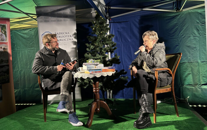 Mężczyzna i kobieta trzymają mikrofony w rękach i siedzą na krzesłach na scenie. Pomiędzy nimi jest stolik ze stosem książek i choinka.