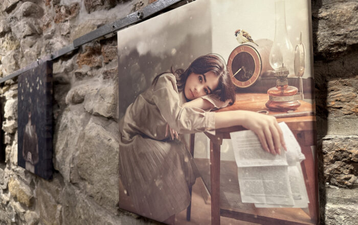 Fotografia na płótnie. Kobieta w długiej beżowej sukience i brązowych włosach trzyma głowę opartą na jednej ręce. Głowa jej leży na stole. W drugiej ręce trzyma kartki papieru. Na stole stoi także lampa naftowa i zegar z ptaszkiem.