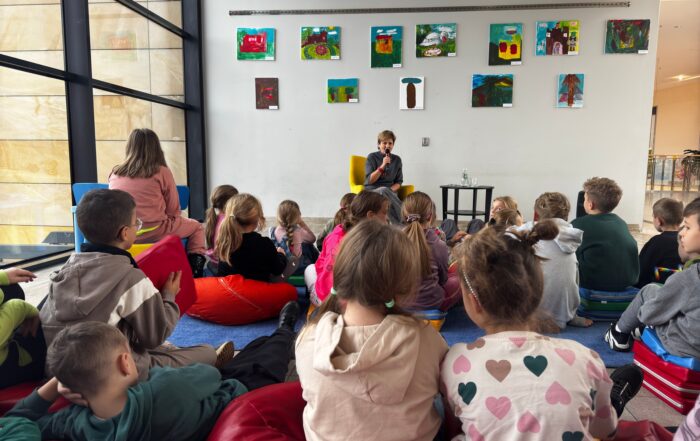 Grupa małych dzieci siedzi na podłodze i słucha kobiety która siedzi z przodu sali i mówi do mikrofonu. Za nią na ścianie wiszą kolorowe obrazy.