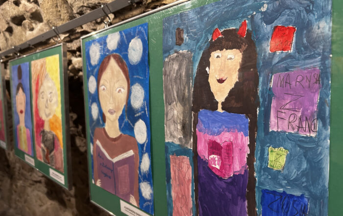 Kolorowe rysunki dzieci przedstawiające kobiety. Wiszą na kamiennej ścianie.